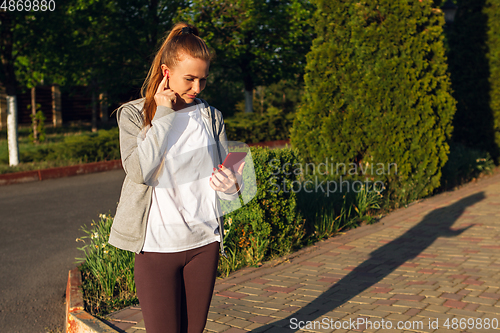 Image of Young female runner, athlete resting after jogging in the city street in sunshine. Beautiful caucasian woman training, listening to music