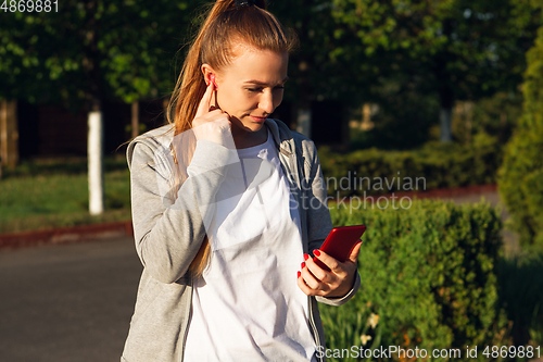 Image of Young female runner, athlete resting after jogging in the city street in sunshine. Beautiful caucasian woman training, listening to music