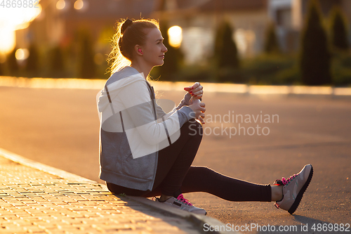 Image of Young female runner, athlete resting after jogging in the city street in sunshine. Beautiful caucasian woman training, listening to music