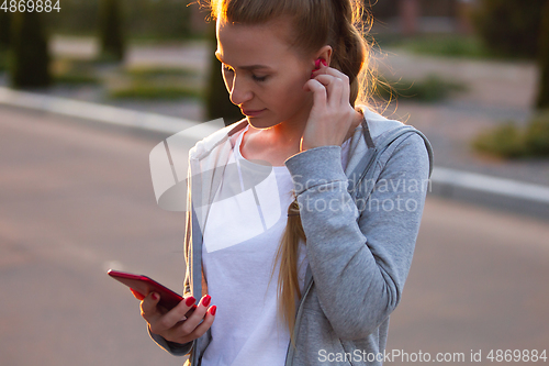 Image of Young female runner, athlete resting after jogging in the city street in sunshine. Beautiful caucasian woman training, listening to music