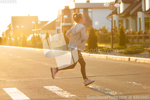 Image of Young female runner, athlete is jogging in the city street in sunshine. Beautiful caucasian woman training, listening to music