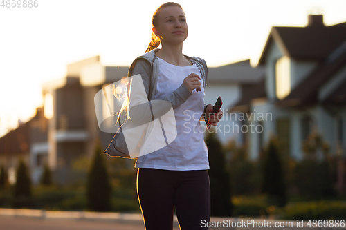 Image of Young female runner, athlete is jogging in the city street in sunshine. Beautiful caucasian woman training, listening to music