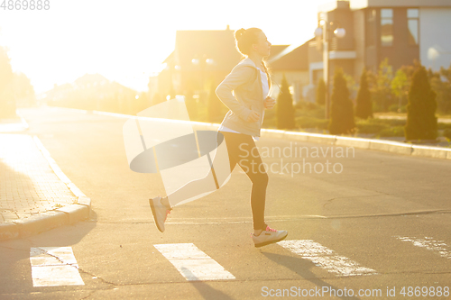 Image of Young female runner, athlete is jogging in the city street in sunshine. Beautiful caucasian woman training, listening to music