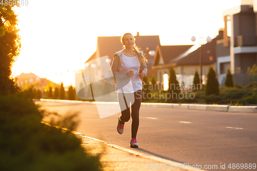 Image of Young female runner, athlete is jogging in the city street in sunshine. Beautiful caucasian woman training, listening to music