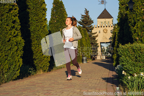 Image of Young female runner, athlete is jogging in the city street in sunshine. Beautiful caucasian woman training, listening to music