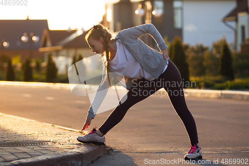 Image of Young female runner, athlete is stretching before jogging in the city street in sunshine. Beautiful caucasian woman training, listening to music