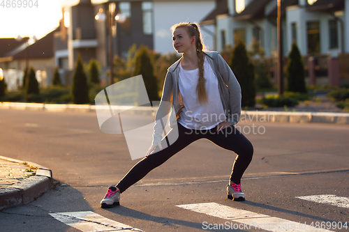 Image of Young female runner, athlete is stretching before jogging in the city street in sunshine. Beautiful caucasian woman training, listening to music