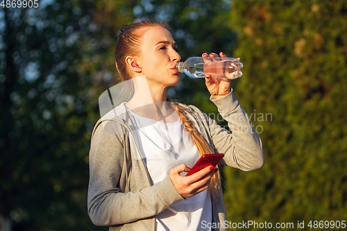 Image of Young female runner, athlete resting after jogging in the city street in sunshine. Beautiful caucasian woman training, listening to music