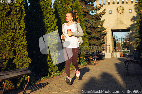 Image of Young female runner, athlete is jogging in the city street in sunshine. Beautiful caucasian woman training, listening to music