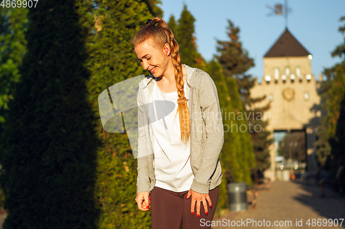 Image of Young female runner, athlete resting after jogging in the city street in sunshine. Beautiful caucasian woman training, listening to music