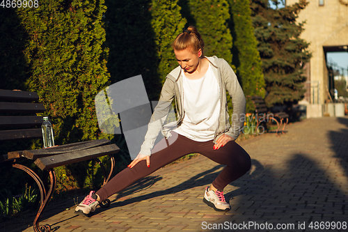 Image of Young female runner, athlete is stretching before jogging in the city street in sunshine. Beautiful caucasian woman training, listening to music