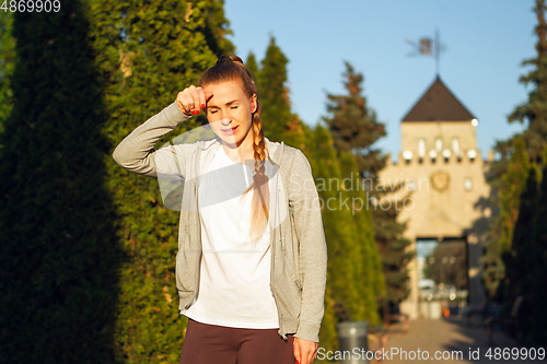 Image of Young female runner, athlete resting after jogging in the city street in sunshine. Beautiful caucasian woman training, listening to music