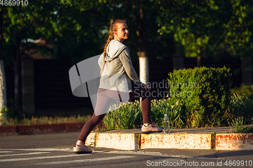 Image of Young female runner, athlete is stretching before jogging in the city street in sunshine. Beautiful caucasian woman training, listening to music