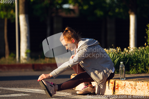 Image of Young female runner, athlete is stretching before jogging in the city street in sunshine. Beautiful caucasian woman training, listening to music