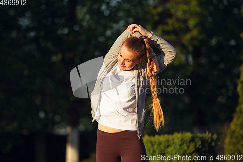 Image of Young female runner, athlete is stretching before jogging in the city street in sunshine. Beautiful caucasian woman training, listening to music