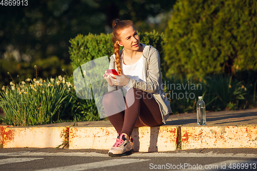 Image of Young female runner, athlete resting after jogging in the city street in sunshine. Beautiful caucasian woman training, listening to music