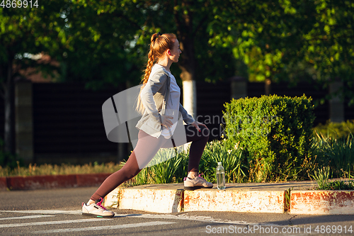 Image of Young female runner, athlete is stretching before jogging in the city street in sunshine. Beautiful caucasian woman training, listening to music