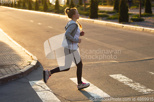 Image of Young female runner, athlete is jogging in the city street in sunshine. Beautiful caucasian woman training, listening to music