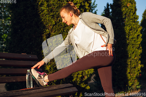 Image of Young female runner, athlete is stretching before jogging in the city street in sunshine. Beautiful caucasian woman training, listening to music