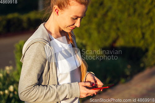 Image of Young female runner, athlete resting after jogging in the city street in sunshine. Beautiful caucasian woman training, listening to music