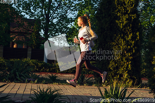 Image of Young female runner, athlete is jogging in the city street in sunshine. Beautiful caucasian woman training, listening to music