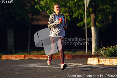 Image of Young female runner, athlete is jogging in the city street in sunshine. Beautiful caucasian woman training, listening to music