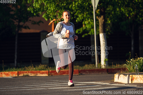 Image of Young female runner, athlete is jogging in the city street in sunshine. Beautiful caucasian woman training, listening to music