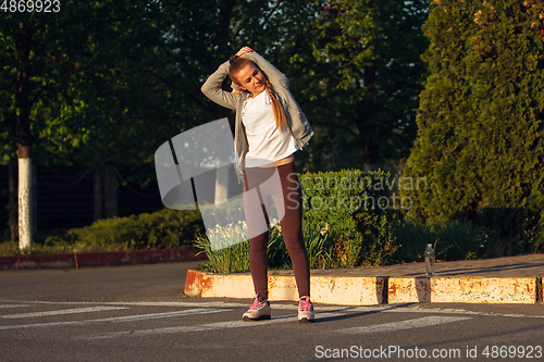 Image of Young female runner, athlete is stretching before jogging in the city street in sunshine. Beautiful caucasian woman training, listening to music