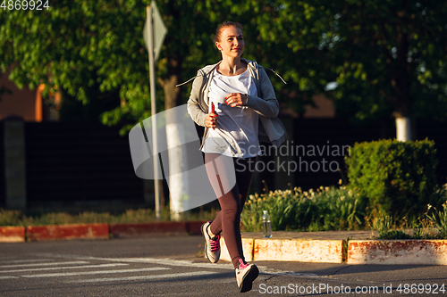 Image of Young female runner, athlete is jogging in the city street in sunshine. Beautiful caucasian woman training, listening to music