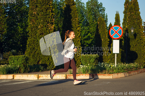 Image of Young female runner, athlete is jogging in the city street in sunshine. Beautiful caucasian woman training, listening to music