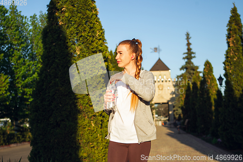 Image of Young female runner, athlete resting after jogging in the city street in sunshine. Beautiful caucasian woman training, listening to music