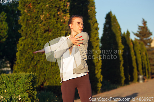 Image of Young female runner, athlete is stretching before jogging in the city street in sunshine. Beautiful caucasian woman training, listening to music