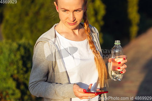 Image of Young female runner, athlete resting after jogging in the city street in sunshine. Beautiful caucasian woman training, listening to music