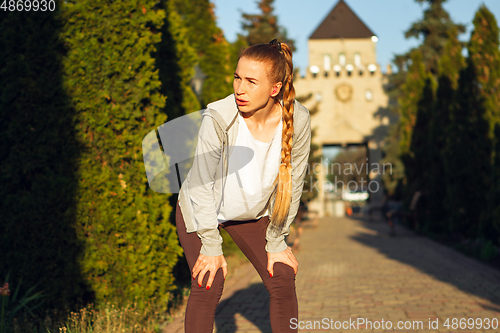 Image of Young female runner, athlete resting after jogging in the city street in sunshine. Beautiful caucasian woman training, listening to music