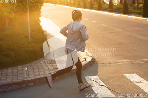 Image of Young female runner, athlete is jogging in the city street in sunshine. Beautiful caucasian woman training, listening to music