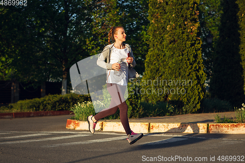 Image of Young female runner, athlete is jogging in the city street in sunshine. Beautiful caucasian woman training, listening to music