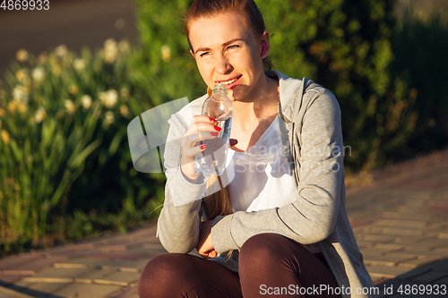 Image of Young female runner, athlete resting after jogging in the city street in sunshine. Beautiful caucasian woman training, listening to music