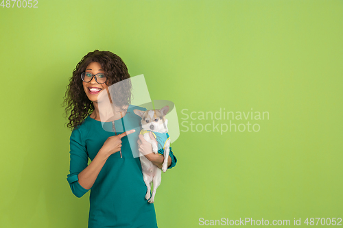 Image of African-american beautiful young woman\'s portrait with little doggy on green studio background, emotional and expressive. Copyspace for ad.