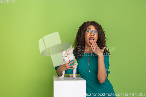 Image of African-american beautiful young woman\'s portrait with little doggy on green studio background, emotional and expressive. Copyspace for ad.