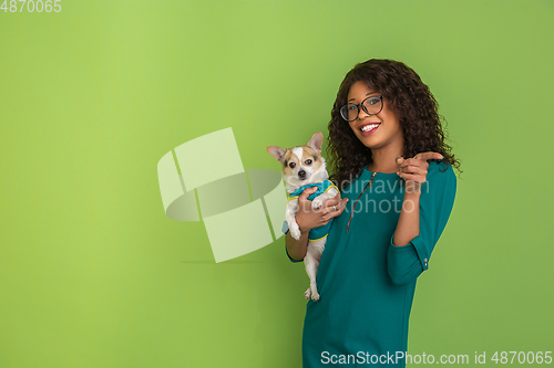 Image of African-american beautiful young woman\'s portrait with little doggy on green studio background, emotional and expressive. Copyspace for ad.