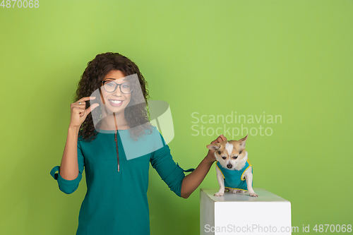 Image of African-american beautiful young woman\'s portrait with little doggy on green studio background, emotional and expressive. Copyspace for ad.