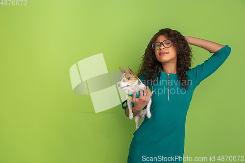 Image of African-american beautiful young woman\'s portrait with little doggy on green studio background, emotional and expressive. Copyspace for ad.