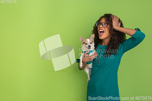 Image of African-american beautiful young woman\'s portrait with little doggy on green studio background, emotional and expressive. Copyspace for ad.