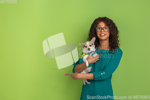 Image of African-american beautiful young woman\'s portrait with little doggy on green studio background, emotional and expressive. Copyspace for ad.