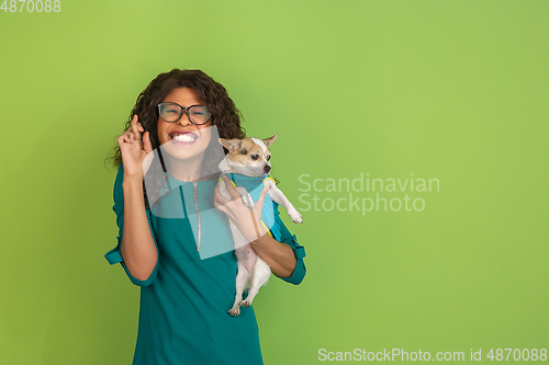 Image of African-american beautiful young woman\'s portrait with little doggy on green studio background, emotional and expressive. Copyspace for ad.