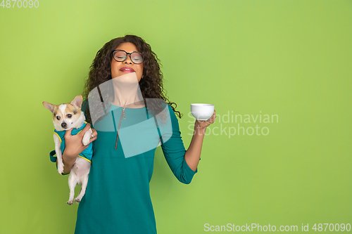 Image of African-american beautiful young woman\'s portrait with little doggy on green studio background, emotional and expressive. Copyspace for ad.