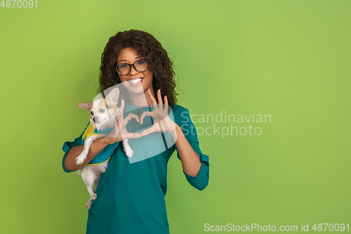 Image of African-american beautiful young woman\'s portrait with little doggy on green studio background, emotional and expressive. Copyspace for ad.