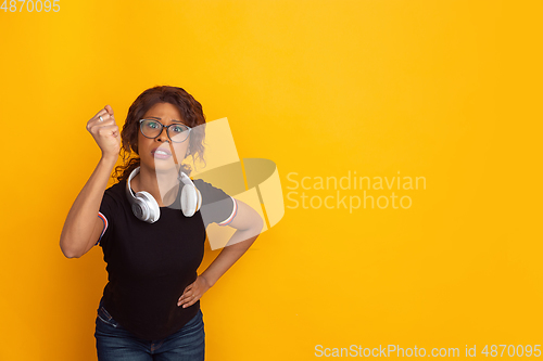 Image of African-american beautiful young woman\'s portrait with wireless headphones on yellow studio background, emotional and expressive. Copyspace for ad.