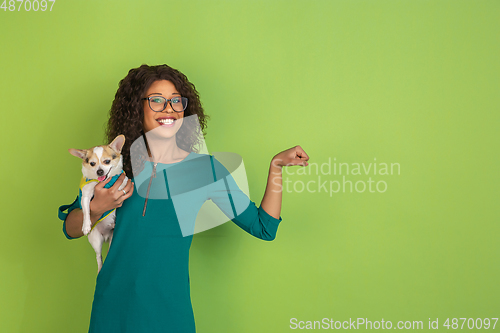Image of African-american beautiful young woman\'s portrait with little doggy on green studio background, emotional and expressive. Copyspace for ad.
