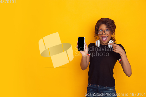 Image of African-american beautiful young woman\'s portrait with wireless headphones on yellow studio background, emotional and expressive. Copyspace for ad.